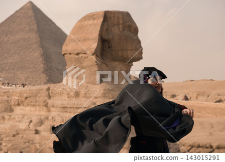 Young woman wearing a dramatic windswept black abaya glances over her shoulder before the Great Sphinx and Pyramid of Giza on the ancient sandy Young woman wearing a dramatic windswept black abaya glances over her shoulder before the Great Sphinx and Pyramid of Giza on the ancient sandy 143015291