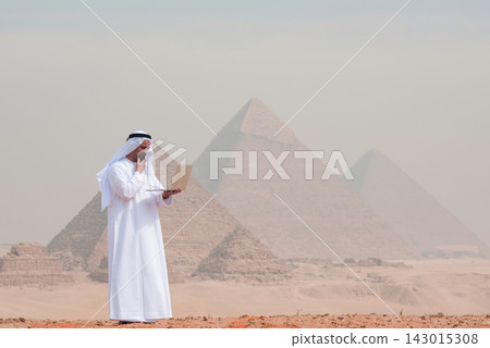 Contemplative arab businessman in traditional white kandura holding laptop outdoors at iconic egyptian landmark blending heritage with modern Contemplative arab businessman in traditional white kandura holding laptop outdoors at iconic egyptian landmark blending heritage with modern 143015308
