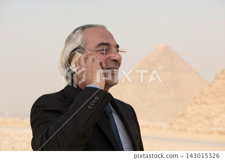 Smiling senior Middle Eastern businessman making a cell phone call with the ancient Giza Pyramids glowing in bokeh behind him ideal for MENA executive Smiling senior Middle Eastern businessman making a cell phone call with the ancient Giza Pyramids glowing in bokeh behind him ideal for MENA executive 143015326