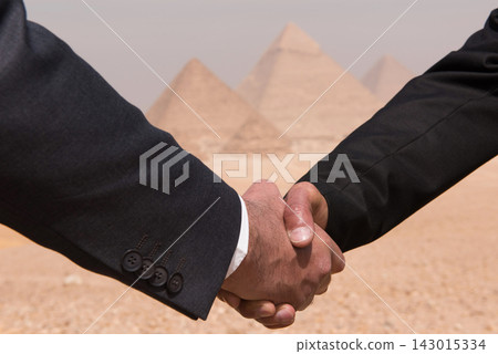 Two business executives in dark suits sealing an international deal with a firm handshake against the iconic Giza pyramids in Egypt ideal for global Two business executives in dark suits sealing an international deal with a firm handshake against the iconic Giza pyramids in Egypt ideal for global 143015334