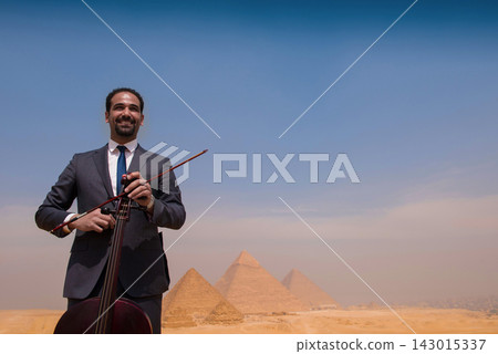 Proud Arab classical musician in formal gray suit smiling while holding cello and bow outdoors before the iconic Giza Pyramids under a vast blue sky 143015337