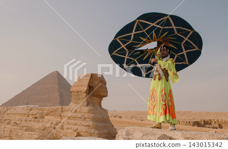 Theatrical folk performer in bold lime green embroidered robe and large teal geometric parasol posing beside Great Sphinx with pyramid of Giza on Theatrical folk performer in bold lime green embroidered robe and large teal geometric parasol posing beside Great Sphinx with pyramid of Giza on 143015342