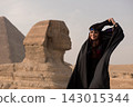 Joyful young Middle Eastern woman in ornate embroidered black folk costume playfully adjusting beaded head wrap with the iconic Great Sphinx and Giza 143015344