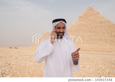 Animated Arab professional in traditional white thobe laughing and pointing during cell phone call with the iconic Giza pyramids rising in the desert Animated Arab professional in traditional white thobe laughing and pointing during cell phone call with the iconic Giza pyramids rising in the desert 143015348