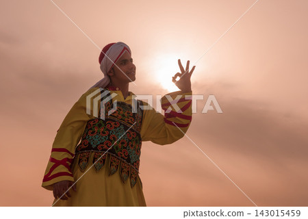 Proud young Middle Eastern man in ornate embroidered Arab folk costume raises OK gesture as sun aligns inside his fingers against a warm golden sunset 143015459