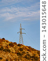Electricity pylon standing on sunlit arid hillside with dry shrubs and golden grasses under blue sky with scattered clouds and circling birds during late afternoon light 143015648