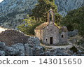 Small stone chapel in mountain ruins with olive trees and dry stone terraces under warm morning sunlight, rocky pathway and weathered masonry in Mediterranean countryside 143015650