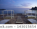 Empty canopy daybeds on sandy beach with centered wooden walkway stretching toward calm sea beneath pastel evening sky and soft clouds in symmetrical composition 143015654