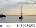 Single sailboat anchored on calm ocean at dusk under pastel evening sky with distant rocky headland and soft horizon light 143015655