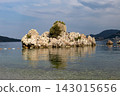 Rocky islet rising from calm shallow sea with reflected sky, pebbly shoreline in foreground, and distant coastline hills and cliffs under partly cloudy bright daylight 143015656