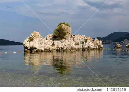 Rocky islet rising from calm shallow sea with reflected sky, pebbly shoreline in foreground, and distant coastline hills and cliffs under partly cloudy bright daylight Rocky islet rising from calm shallow sea with reflected sky, pebbly shoreline in foreground, and distant coastline hills and cliffs under partly cloudy bright daylight 143015656