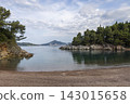 Calm pebble beach cove between rocky pine covered cliffs, with distant tree island and overcast sky, glassy sea reflections and curving shoreline 143015658