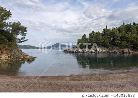Calm pebble beach cove between rocky pine covered cliffs, with distant tree island and overcast sky, glassy sea reflections and curving shoreline Calm pebble beach cove between rocky pine covered cliffs, with distant tree island and overcast sky, glassy sea reflections and curving shoreline 143015658