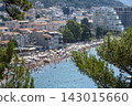 Crowded Mediterranean beach with colorful umbrellas and coastal town skyline viewed from pine hillside in bright summer daylight 143015660