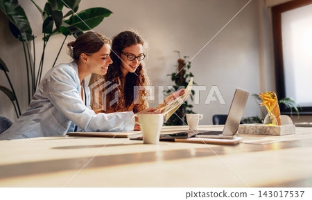 Creative business partners study a printed diagram over coffee at a sunlit table surrounded by lush green office plants Creative business partners study a printed diagram over coffee at a sunlit table surrounded by lush green office plants 143017537