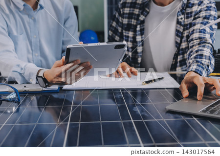 Two Asian engineers wearing hardhats and safety vests discussing solar panel installation and renewable energy project in the office. Two Asian engineers wearing hardhats and safety vests discussing solar panel installation and renewable energy project in the office. 143017564