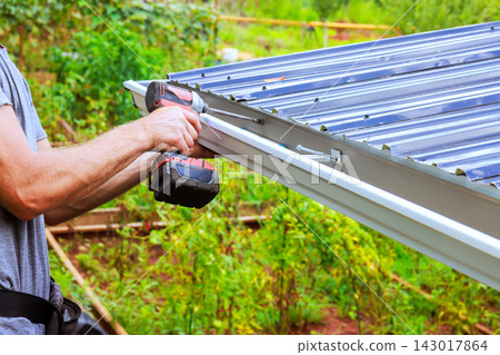 Man works with power drill to install metal roof gutter in garden setting. Man works with power drill to install metal roof gutter in garden setting. 143017864