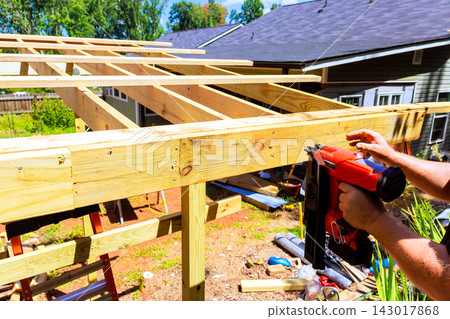 Builder is using air nail gun to secure wooden beams in backyard with tools, materials are scattered around. Builder is using air nail gun to secure wooden beams in backyard with tools, materials are scattered around. 143017868