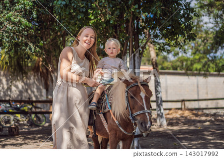 Mother and little boy share a happy pony riding experience. Family bonding through outdoor riding activities. Mother and little boy share a happy pony riding experience. Family bonding through outdoor riding activities. 143017992