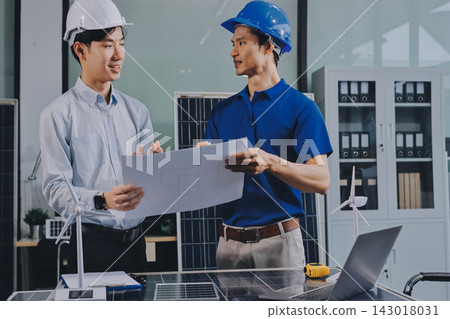 Two Asian engineers wearing hardhats and safety vests discussing solar panel installation and renewable energy project in the office. Two Asian engineers wearing hardhats and safety vests discussing solar panel installation and renewable energy project in the office. 143018031