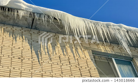 Long, powerful icicles hang from the snow-covered edge of the roof of a brick residential building. Long, powerful icicles hang from the snow-covered edge of the roof of a brick residential building. 143018666