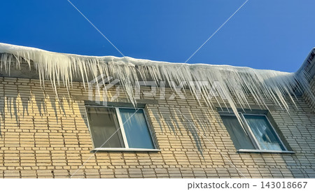 Long, powerful icicles hang from the snow-covered edge of the roof of a brick residential building. 143018667