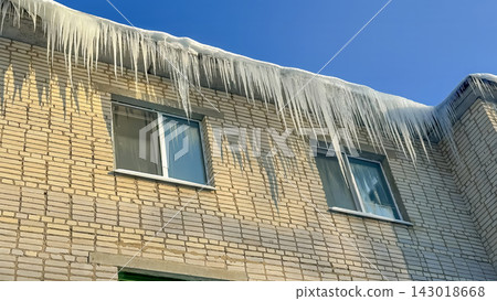Long, powerful icicles hang from the snow-covered edge of the roof of a brick residential building. Long, powerful icicles hang from the snow-covered edge of the roof of a brick residential building. 143018668