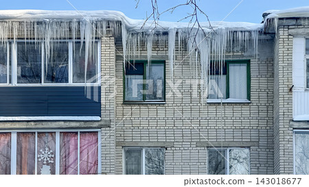 Long, powerful icicles hang from the snow-covered edge of the roof of a brick residential building. Long, powerful icicles hang from the snow-covered edge of the roof of a brick residential building. 143018677