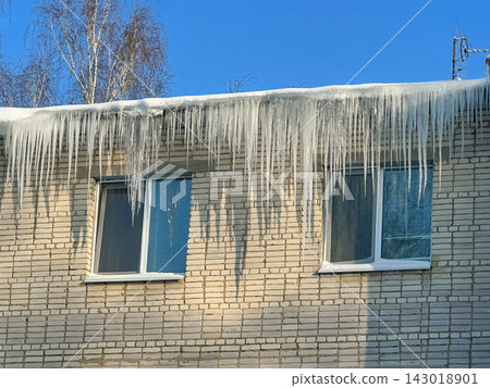 Long, powerful icicles hang from the snow-covered edge of the roof of a brick residential building. Long, powerful icicles hang from the snow-covered edge of the roof of a brick residential building. 143018901