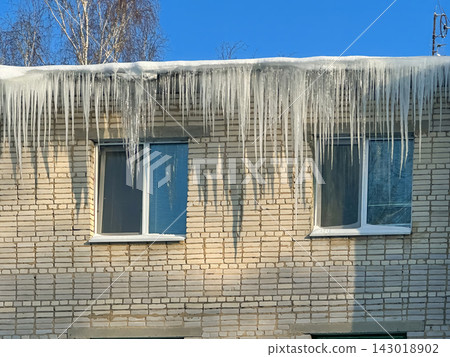 Long, powerful icicles hang from the snow-covered edge of the roof of a brick residential building. 143018902