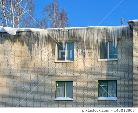 Long, powerful icicles hang from the snow-covered edge of the roof of a brick residential building. Long, powerful icicles hang from the snow-covered edge of the roof of a brick residential building. 143018903