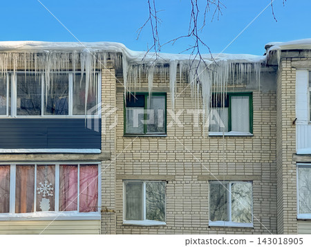 Long, powerful icicles hang from the snow-covered edge of the roof of a brick residential building. Long, powerful icicles hang from the snow-covered edge of the roof of a brick residential building. 143018905
