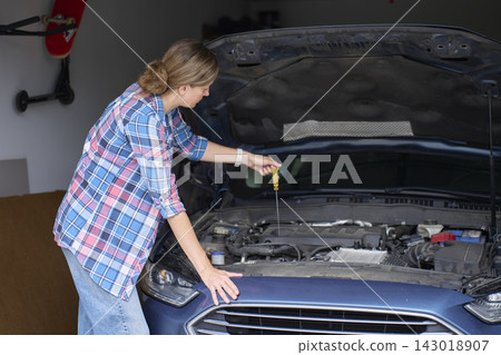 Woman using dipstick to check engine oil under open car hood in home garage 143018907