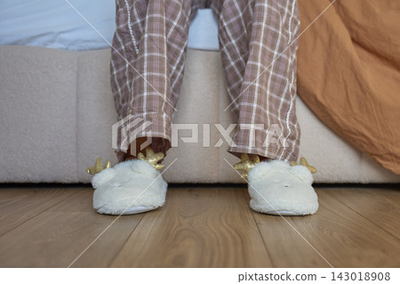 Person wearing fluffy white slippers with pajama pants on wooden floor beside bed 143018908