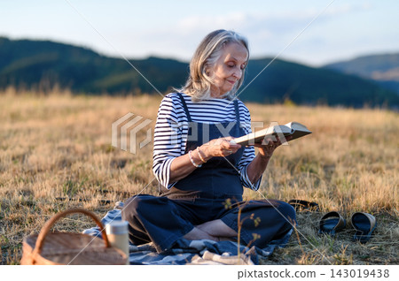 Older woman enjoying peaceful solo picnic and reading in countryside. Older woman enjoying peaceful solo picnic and reading in countryside. 143019438