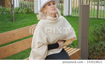 Mature woman holds abdomen while seated on garden bench in cream clothing. Older adult wears elegant white hat and appears worried, presenting reflective senior lifestyle portrait outdoors. Mature woman holds abdomen while seated on garden bench in cream clothing. Older adult wears elegant white hat and appears worried, presenting reflective senior lifestyle portrait outdoors. 143019621