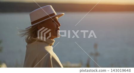 Woman in white hat looks toward tranquil water and glowing sunset. Older woman wears pale coat and scarf, her serene profile expressing quiet reflection beside softly blurred horizon. 143019648