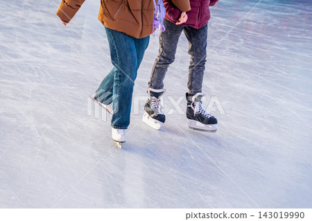 Two People Ice Skating Together on Winter Rink. Cropped view of two people skating together across outdoor ice rink. Ice skates, casual winter clothing, frozen surface illustrate seasonal recreation 143019990