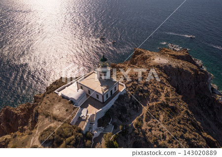 Elevated aerial view of Akrotiri Lighthouse on the rugged southwestern coast of Santorini, Greece, surrounded by shimmering Aegean Sea and rocky shoreline. Elevated aerial view of Akrotiri Lighthouse on the rugged southwestern coast of Santorini, Greece, surrounded by shimmering Aegean Sea and rocky shoreline. 143020889