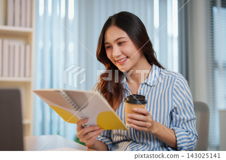 Smiling Asian woman holding coffee cup and reading notebook at office desk 143025141