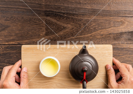 Top view of hands holding wooden tray with clay teapot and tea cup on dark wooden background with copy space 143025858