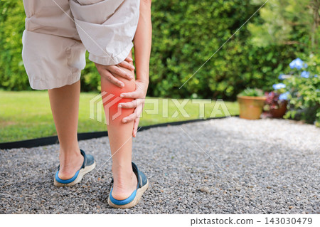 Close up of person standing holding bare calf with red highlighted area indicating severe leg pain, muscle cramp, strain, or sports injury. Healthcare, physical therapy, and medical concept outdoors. Close up of person standing holding bare calf with red highlighted area indicating severe leg pain, muscle cramp, strain, or sports injury. Healthcare, physical therapy, and medical concept outdoors. 143030479