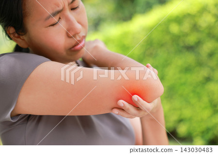 Stressed young Asian woman holding her elbow with red highlighted area indicating severe joint pain, arthritis, muscle sprain or sports injury. Healthcare, physical therapy, and medical care concept. Stressed young Asian woman holding her elbow with red highlighted area indicating severe joint pain, arthritis, muscle sprain or sports injury. Healthcare, physical therapy, and medical care concept. 143030483
