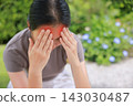 Stressed young Asian woman sitting outdoors touching her head with red highlighted area indicating severe headache, migraine, or brain pain. Concept of sickness, healthcare, tension and mental health. 143030487