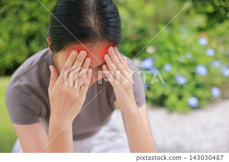Stressed young Asian woman sitting outdoors touching her head with red highlighted area indicating severe headache, migraine, or brain pain. Concept of sickness, healthcare, tension and mental health. Stressed young Asian woman sitting outdoors touching her head with red highlighted area indicating severe headache, migraine, or brain pain. Concept of sickness, healthcare, tension and mental health. 143030487
