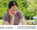 Unhappy young Asian woman sitting outdoors touching her cheek with a red highlighted area indicating severe toothache, dental pain, gum disease, or jaw problem. Healthcare, dentist, medical concept. 143030488
