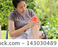 Stressed young Asian woman sitting on a chair touching her bare knee with red highlighted area indicating severe joint pain, arthritis, muscle ache or sports injury. Healthcare, medical concept. 143030489