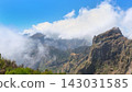 Mountain peaks covered by low clouds under a bright blue sky with rugged cliffs and green vegetation in a scenic natural landscape, Madeira, Portugal. 143031585