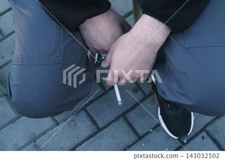 Person sitting on a stone floor holding a lit cigarette in their hand Person sitting on a stone floor holding a lit cigarette in their hand 143032502