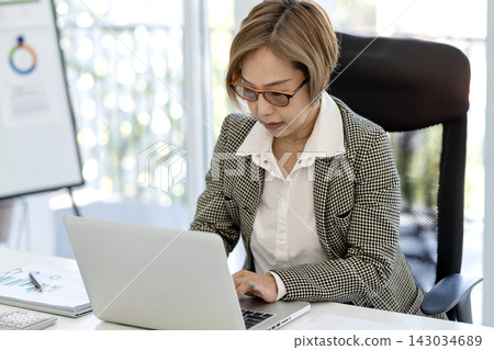Senior Asian Businesswoman Working on Laptop at Modern Office Desk with Financial Documents Senior Asian Businesswoman Working on Laptop at Modern Office Desk with Financial Documents 143034689